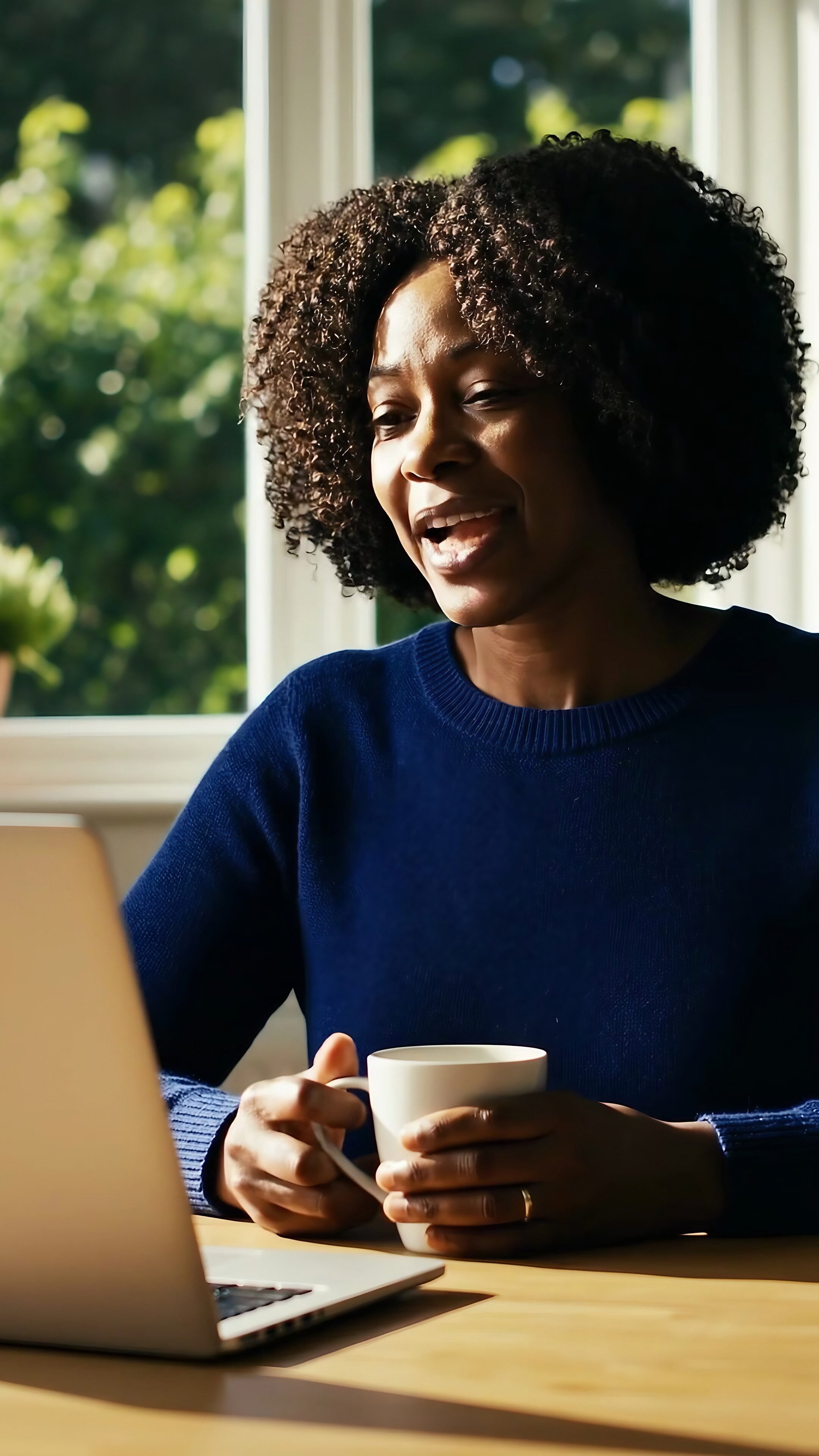A woman at her kitchen table by a sunny window, talking with her provider on a laptop, a mug of tea in her hands.